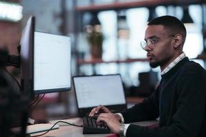 ung afrikansk amerikan med glasögon Sammanträde i en modern coworking kontor och arbetssätt på en dator foto