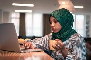 afrikansk muslim företag kvinna bär en grön hijab dricka te medan arbetssätt på bärbar dator dator i avslappning område på modern öppen planen börja kontor. foto