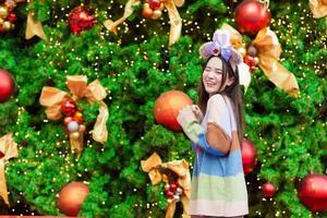 en vacker asiatisk kvinna i en färgglad tröja står leende och glad framför julgranen med bokeh som bakgrund i temat för att fira jul och gott nytt år foto