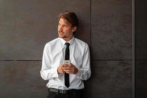 börja affärsman i en vit skjorta med en svart slips använder sig av smartphone medan stående i främre av grå vägg under ha sönder från arbete utanför foto
