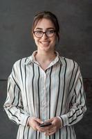 börja affärskvinna i skjorta med en glasögon använder sig av smartphone medan stående i främre av grå vägg under ha sönder från arbete utanför foto
