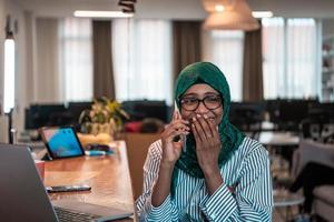 leende ung kvinna prata på smartphone arbete på bärbar dator se i distans tänkande. Lycklig afrikansk amerikan kund klient använda sig av modern gadgetar mobiltelefon och dator på Hem kontor. foto