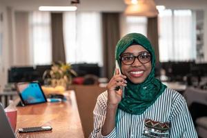 leende ung kvinna prata på smartphone arbete på bärbar dator se i distans tänkande. Lycklig afrikansk amerikan kund klient använda sig av modern gadgetar mobiltelefon och dator på Hem kontor. foto