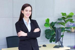 professionell skön asiatisk arbetssätt kvinna vem har lång hår bär svart formell kostym med blå skjorta medan hon ärm korsning och leende lyckligt på de vägg i de kontor. foto