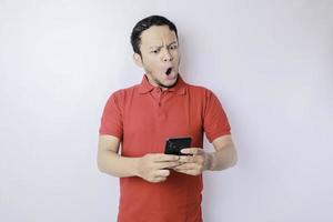 överraskad asiatisk man bär röd t-shirt pekande på hans smartphone, isolerat förbi vit bakgrund foto