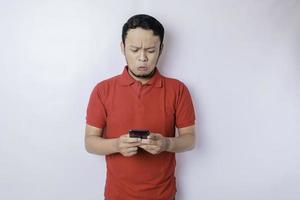 en missnöjd ung asiatisk man utseende missnöjd bär röd t-shirt irriterad ansikte uttryck innehav hans telefon foto