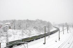 en lång tåg av frakt bilar är rör på sig längs de järnväg Spår. järnväg landskap i vinter- efter snöfall foto