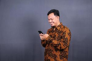 porträtt av asiatisk ung man bär batik använder sig av mobiltelefon på grå bakgrund foto