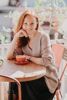 vertikal bild av en glad rävkvinna klädd i modekläder, dricker kaffe, poserar på uteservering, njuter av fritiden, har ett trevligt leende på läpparna, håller handen under hakan. livsstilskoncept foto