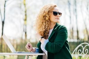 ung vacker kvinna med fluffigt blont hår bär solglasögon sitter på i solen framför laptop översätta texter tittar runt och lägger märke till sin bästa vän som njuter av soligt väder på gården foto