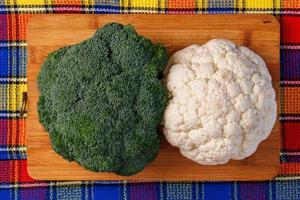 en huvud av broccoli och en huvud av blomkål lögn på en skärande styrelse på de tabell med färgrik handduk under foto