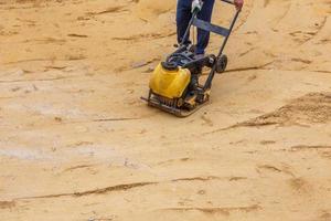 arbetstagare i använda sig av vibrerande tallrik komprimator för kompaktering sand under väg konstruktion. foto