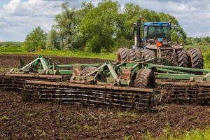 blå traktor med dubbel- hjul dragande skiva harv med vält korg på varm solig dag foto