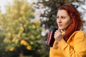 ung rödhårig kvinna drycker kaffe från återanvändbar termo mugg. njut av höst solig dag i natur. foto