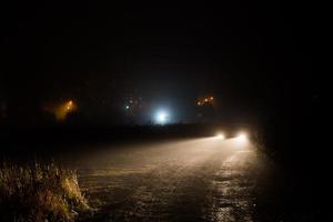 bil strålkastare koner i natt dimma på fält Bakom stad foto