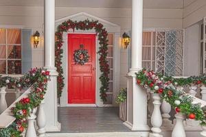jul veranda dekoration aning. hus ingång med röd dörr dekorerad för högtider. röd och grön krans krans av gran träd grenar och lampor på räcke. jul eve på Hem foto