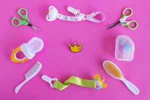 platt lägga på bebis vård objekt - sax, hårborstar, nappar, napp hållare, nasal aspirator och nappare- på rosa bakgrund med liten gul krona för liten prinsessa på födelsedag. foto