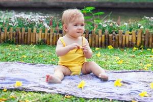 förtjusande liten caucasian flicka i gul klänning är spelar med gul blommor på en pläd. picknick på en sommar solig dag i parkera. foto