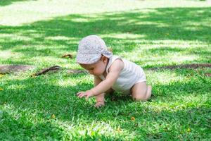 livsstil porträtt av liten bebis flicka i vit Kläder och panama. barn försöker till krypa på gräs i parkera på en solig sommar dag. foto