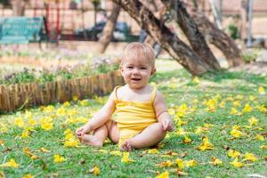 livsstil porträtt av söt bebis flicka med blond hår på gräs med gul blommor. sommar solig dag i parkera. foto