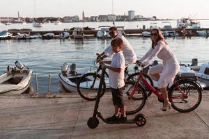 Lycklig familj njuter en skön morgon- förbi de hav tillsammans, föräldrar ridning en cykel och deras son ridning ett elektrisk skoter. selektiv fokus foto