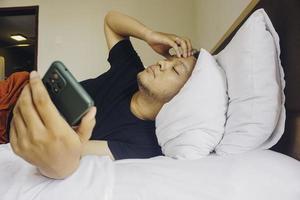 trött ung man kontroll på hans telefon medan liggande i säng foto