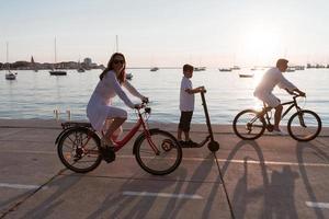 Lycklig familj njuter en skön morgon- förbi de hav tillsammans, föräldrar ridning en cykel och deras son ridning ett elektrisk skoter. selektiv fokus foto