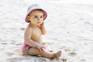 söt liten bebis flicka i rosa simning trunkar och panama Sammanträde på sand på strand. foto