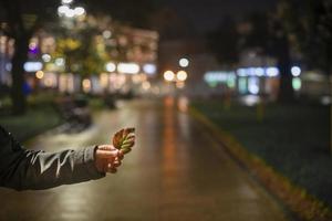 en hand innehar ett höst blad mot de bakgrund av bokeh stad lampor. natt urban landskap, färgad lampor reflekterad i de våt asfalt. de lampor av en regnig natt i de höst stad foto