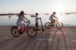 Lycklig familj njuter en skön morgon- förbi de hav tillsammans, föräldrar ridning en cykel och deras son ridning ett elektrisk skoter. selektiv fokus foto