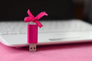 lysande rosa uSB blixt minne kort med en rosa rosett lögner på en filt av mjuk och hårig ljus rosa skinna tyg bredvid till en vit bärbar dator. klassisk kvinna gåva design för en minne kort foto