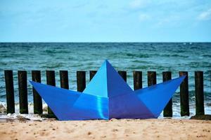 origami blå papper båt på sandig strand för design, blå leksak papper fartyg förbi hav. skön horisont foto