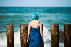 blåhårig kvinna performancekonstnär i blå klänning stående på stranden med pensel, bakifrån foto