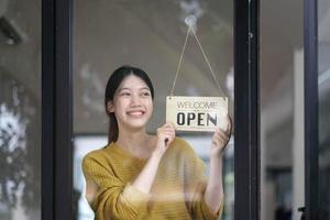 skott av leende asiatisk ung sme små företag ägare bär förkläde och stående vit öppen singnear kaffe affär dörr, asiatisk företag kvinna Barista Kafé ägare sme entreprenör säljare begrepp foto