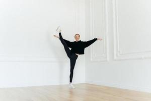 underbar smidig tjej står på ett ben, gör stretchövningar, bär svarta sportkläder, balanserar inomhus, har en nöjd look. sportig kvinna utövar yoga. människor, wellness och fitness koncept foto