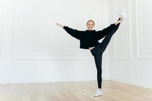 leende sportig kvinna står på ett ben i gymmet, visar skön fysisk spänst, har smal kroppsform, klädd i svart huvtröja och leggings, bär vita sneakers. människor och balans foto