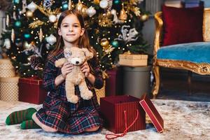 liten bedårande flicka sitter på golvet nära dekorerade nyårsträd, slår in present, är glad över att få nalle från jultomten. litet sött barn får present på julen. mirakel och semester koncept foto