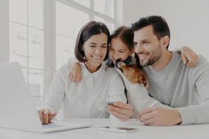 foto av lilla dotter omfamnar sin mamma och pappa, leker med stamtavla, sitta vid ett vitt skrivbord nära datorn, hålla plastkort, kontrollera balansen, posera i det vita rummet. familj, modern teknik