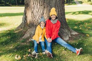 vacker hona bär stickad mössa och swetaer sitter tillsammans med sin lilla dotter nära trädet, vila utomhus i en vacker park, njut av frisk höstluft, känner dig avslappnad. människor, avkoppling foto