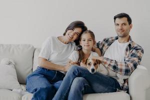 porträtt av tillgiven familj gosa och sitta tillsammans vid soffan i vardagsrummet, byta hem, ha glada uttryck. far, mor, dotter och hund poserar för att göra porträtt, spendera god tid foto