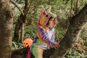 liten flicka i en regnbåge enhörning kostym kigurumi är Sammanträde på en träd med pumpa korg för sötsaker mot de bakgrund av en skog. halloween begrepp. Plats för text. hög kvalitet Foto