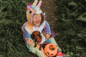 liten skrattande flicka och en dvärg- tax i halloween kostymer med en pumpa korg för sötsaker utomhus. en flicka i en regnbåge enhörning kigurumi kostym, en hund i en klänning med en full kjol. topp se foto