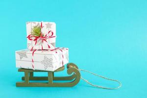 två gåva lådor insvept av vit och grå papper med röd vit band, dekorerad av gran gren på en grön trä- släde på en ljus blå bakgrund. jul och ny år begrepp med kopia Plats. foto