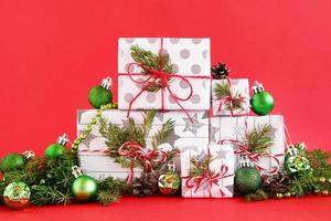 jul gåva lådor insvept av vit-grå papper med röd vit band på en röd bakgrund, dekorerad av gran grenar, tall koner och skinande grön jul dekorationer. foto