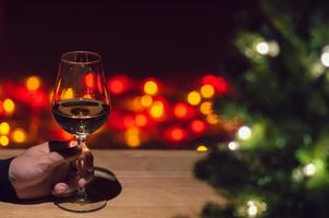 hand toasting en glas av reste sig vin på trä- tabell med jul träd och färgrik bokeh ljus bakgrund. foto