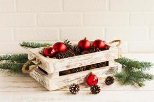 en vit handgjort trä- låda med röd jul bollar och grenar av gran med koner på en trä- tabell. foto