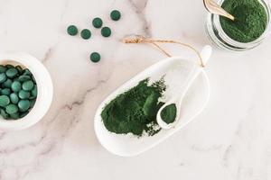 organisk grön spirulina pulver och grön alger tabletter i en vit skål och sked på en marmor tabell. topp se. mat källa av vitamin protein. foto