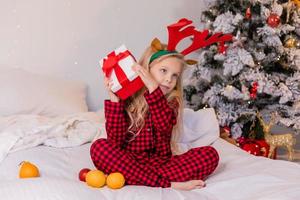 Lycklig liten blond flicka på Hem i pyjamas för jul sortering presenterar och spelar med jul leksaker foto