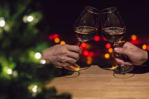 två händer toasting glasögon av reste sig vin på trä- tabell med jul träd och färgrik bokeh ljus bakgrund. foto