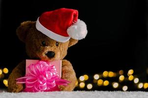 brun teddy Björn bär santa claus hatt innehav partiell fokus av jul gåva låda och bokeh lampor bakgrund. foto